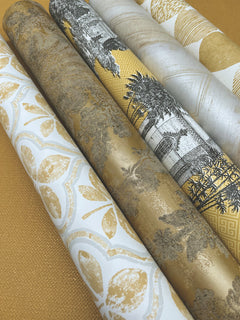 Five rolls of Ronald Redding Charleston Damask Wallpaper by York Wallcoverings featuring various botanical and architectural patterns in gold, white, and black tones.