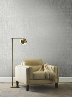An armchair with a throw blanket and a floor lamp positioned in front of a wall featuring Ronald Redding Liquid Metal Wallpaper with a textured silver finish.