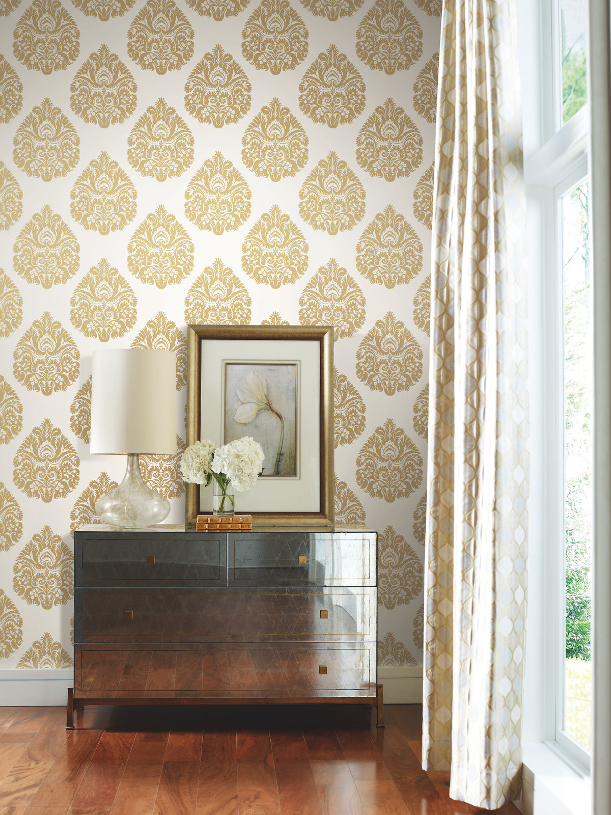 Ronald Redding Teardrop Damask Wallpaper with a repeating gold damask teardrop pattern on a white background, installed behind a dresser with a lamp and framed floral print.
