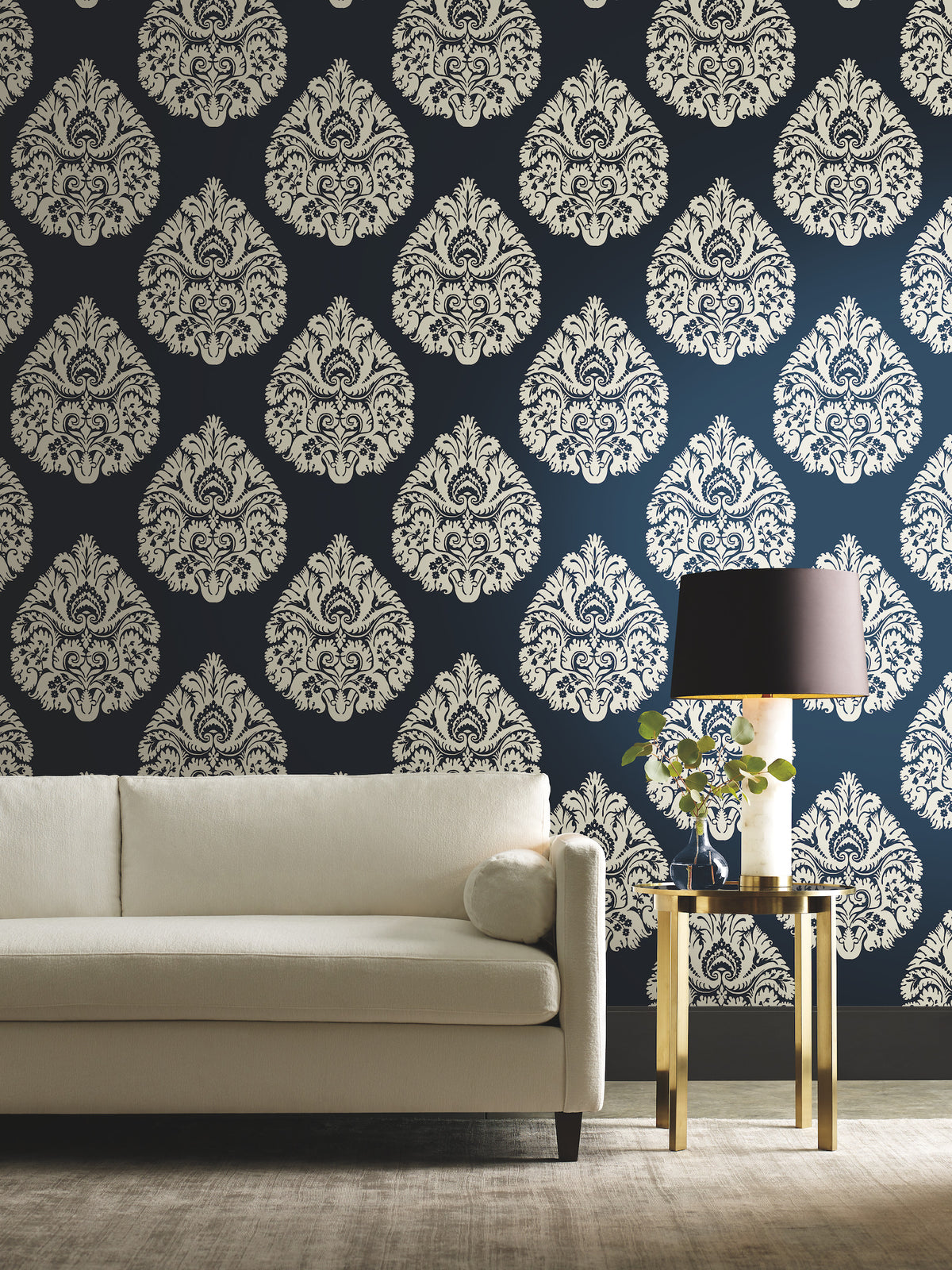 Cream-colored Ronald Redding Teardrop Damask Wallpaper with a repeating teardrop pattern on a dark blue background behind a white sofa and gold side table.