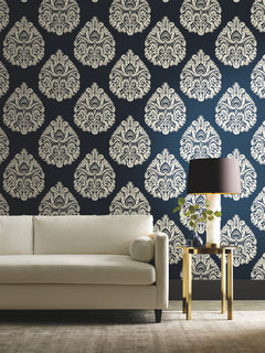 Cream-colored Ronald Redding Teardrop Damask Wallpaper with a repeating teardrop pattern on a dark blue background behind a white sofa and gold side table.