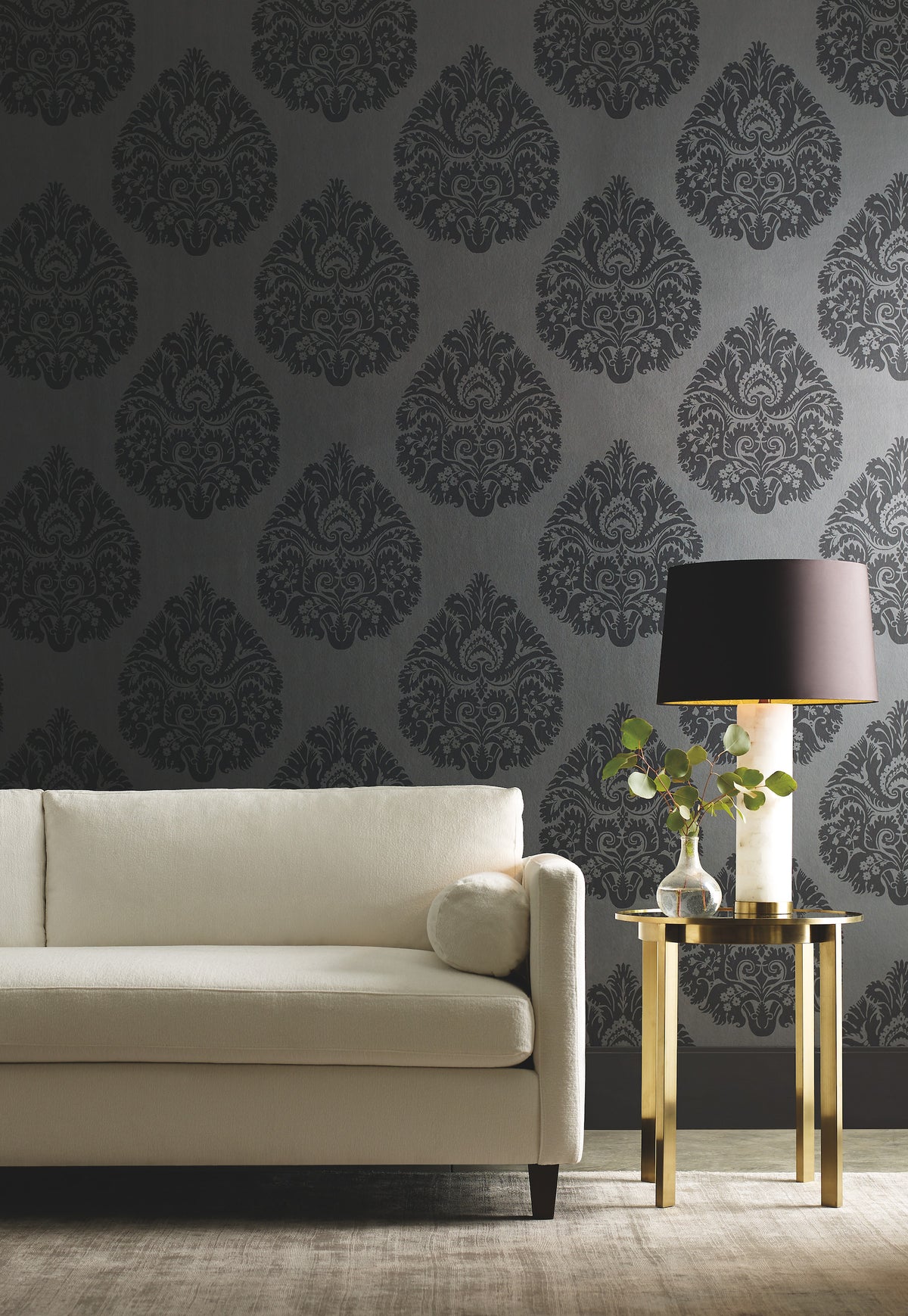 Ronald Redding Teardrop Damask Wallpaper in dark gray with a repeating teardrop pattern behind a cream-colored sofa and a gold side table with a lamp.