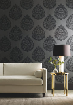 Ronald Redding Teardrop Damask Wallpaper in dark gray with a repeating teardrop pattern behind a cream-colored sofa and a gold side table with a lamp.