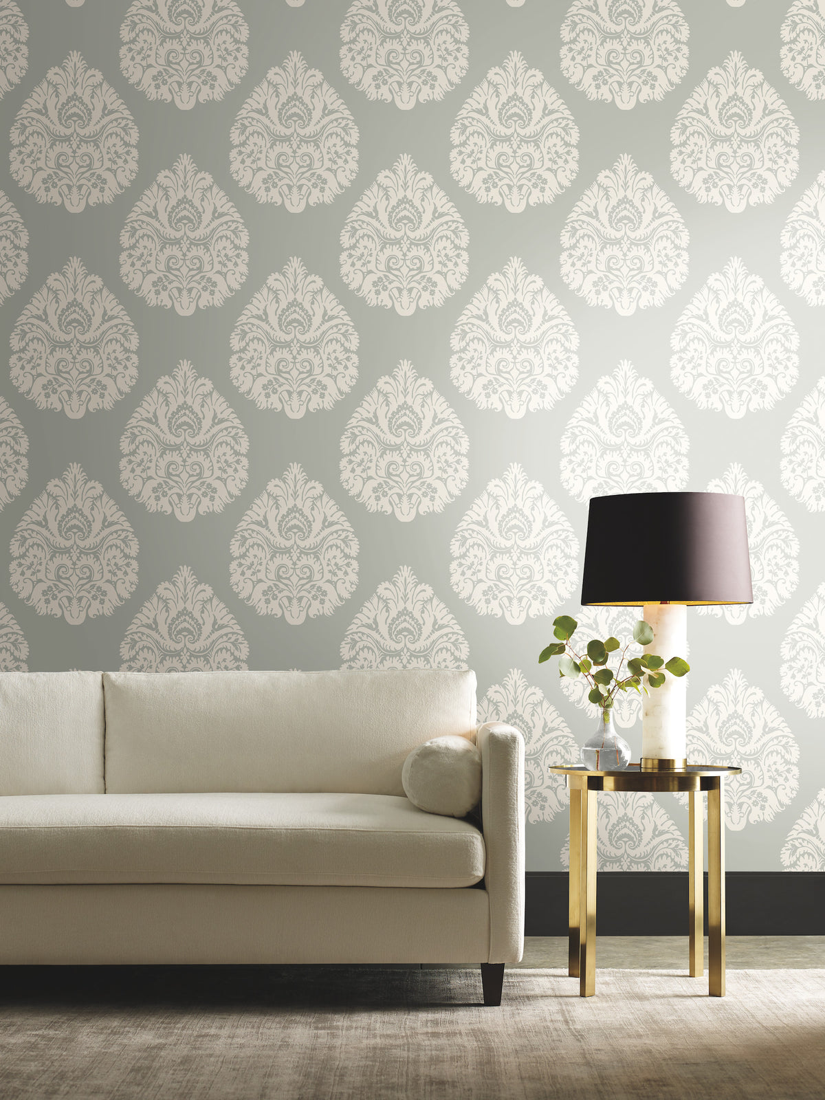 Ronald Redding Teardrop Damask Wallpaper in white features a teardrop pattern on a sage green background, displayed behind a neutral sofa and a gold side table with a lamp.