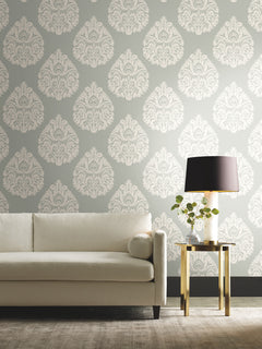 Ronald Redding Teardrop Damask Wallpaper in white features a teardrop pattern on a sage green background, displayed behind a neutral sofa and a gold side table with a lamp.