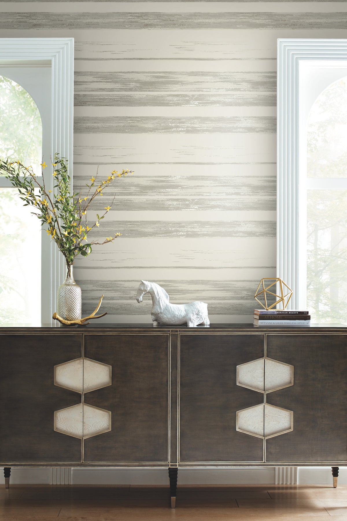 Ronald Redding Horizontal Dry Brush Wallpaper with a neutral-toned texture behind a dark sideboard holding a vase of yellow flowers, a horse figurine, and a stack of books.