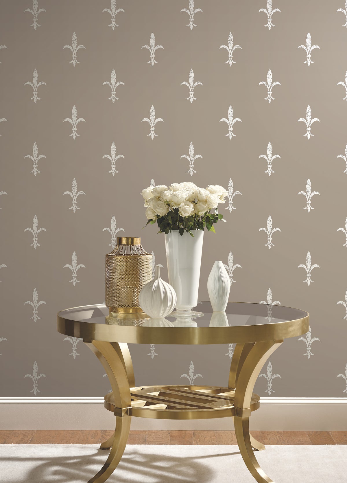 A gold metal table with a vase of white roses and two decorative vessels set against Ronald Redding Fleur De Lis Wallpaper by York Wallcoverings, featuring a repeating white fleur-de-lis pattern on a taupe background.
