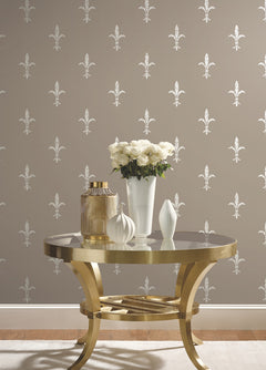 A gold metal table with a vase of white roses and two decorative vessels set against Ronald Redding Fleur De Lis Wallpaper by York Wallcoverings, featuring a repeating white fleur-de-lis pattern on a taupe background.