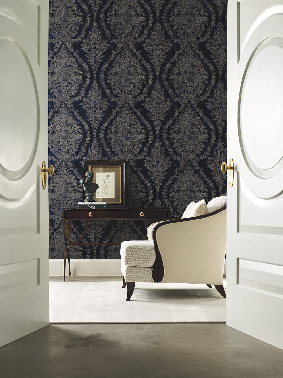 A room viewed through open double doors, featuring dark blue Ronald Redding Charleston Damask Wallpaper by York Wallcoverings, a dark wood console table, and a cream-colored armchair.