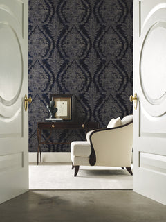 A room viewed through open double doors, featuring dark blue Ronald Redding Charleston Damask Wallpaper by York Wallcoverings, a dark wood console table, and a cream-colored armchair.