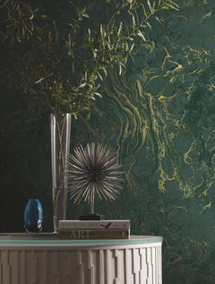 Ronald Redding Polished Marble Wallpaper in dark green with gold veining, displayed behind a glass vase with branches, a spiky metal sculpture, and two books on a white console table.