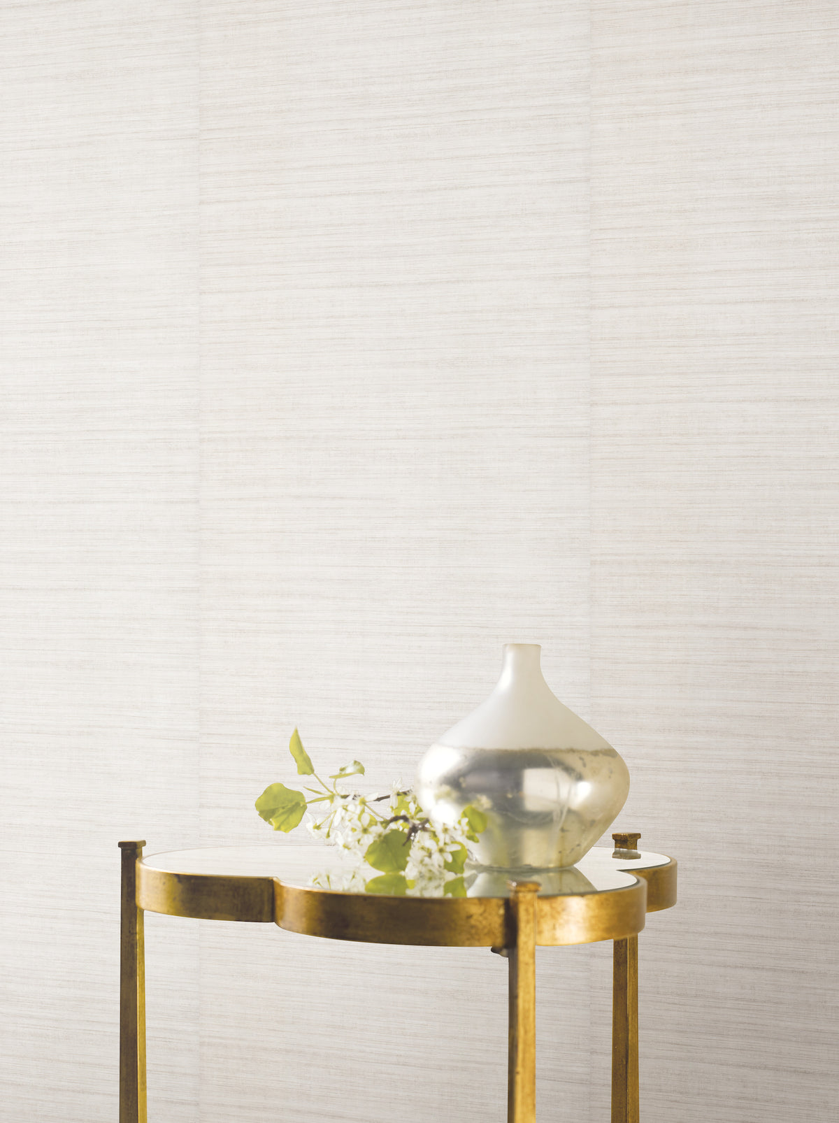 A small gold table holds a reflective silver vase with green leafy branches, set against a textured wall adorned with Ronald Redding Silk Elegance Wallpaper by York Wallcoverings.