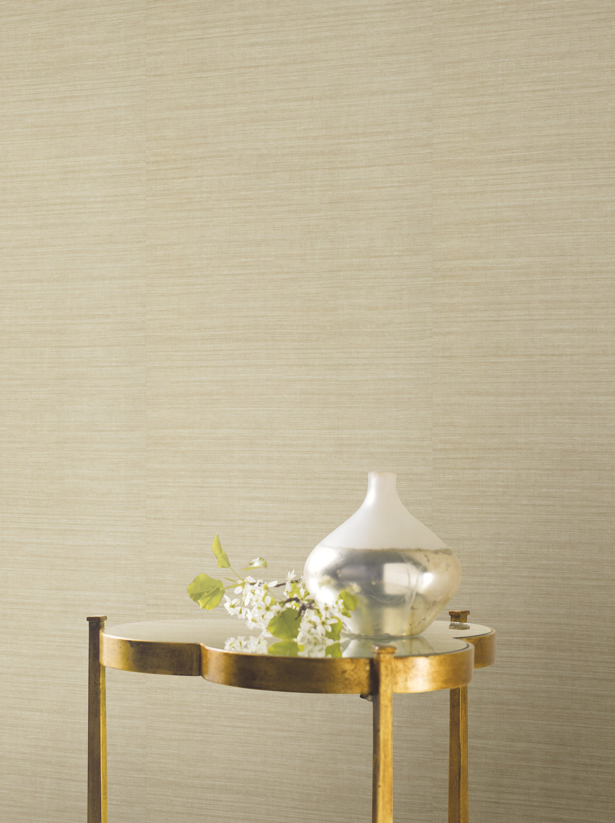 Ronald Redding Silk Elegance Wallpaper in beige, featuring a fine, horizontal fabric-like texture, shown behind a small metallic table holding a decorative vase and a small branch.