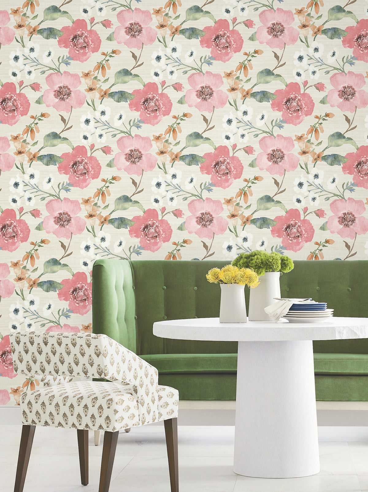 A white round table with yellow flowers in a vase, a white upholstered chair, and a green bench are arranged in front of a wall adorned with York Wallcoverings’ Evelyn Floral Wallpaper.