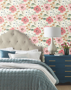 A bedroom featuring the Evelyn Floral Wallpaper by York Wallcoverings, a tufted beige headboard, a blue quilted bedspread, a blue nightstand, a white lamp, and a vase of tulips. Pillows are neatly arranged on the bed.