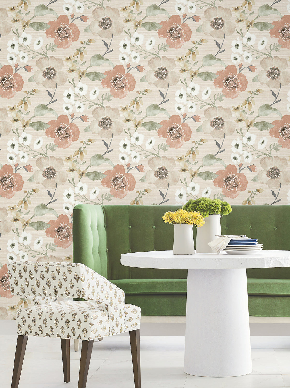 An upholstered green bench, a round white table adorned with two vases of yellow flowers, and a patterned chair are arranged in front of the York Wallcoverings “Evelyn Floral Wallpaper,” which features peach, white, and green blooms.
