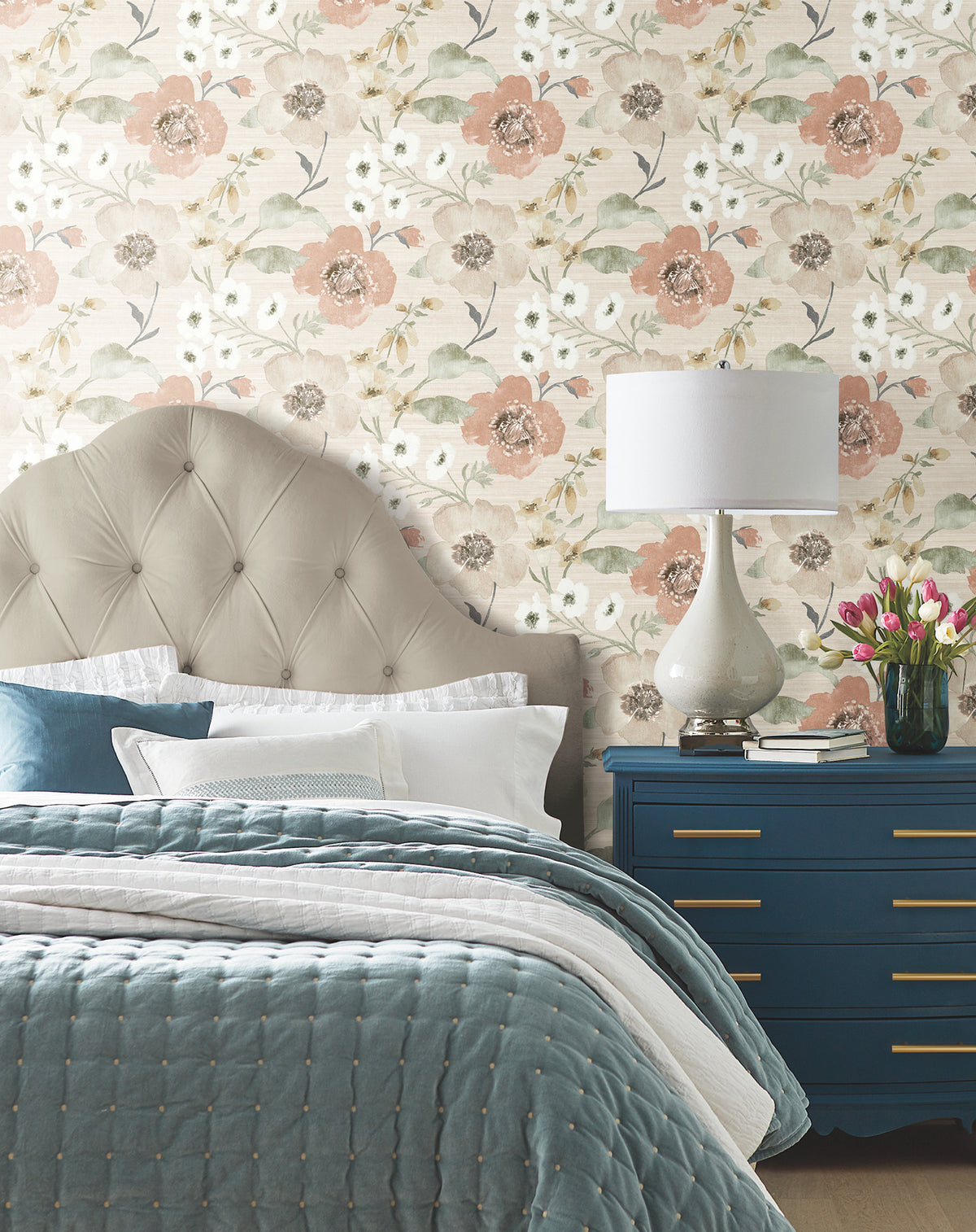 Bedroom with a tufted headboard, blue quilted bedding, a navy dresser with gold handles, a white lamp, and a vase of pink tulips. The wall features York Wallcoverings’ Evelyn Floral Wallpaper in pink, white, and green tones.