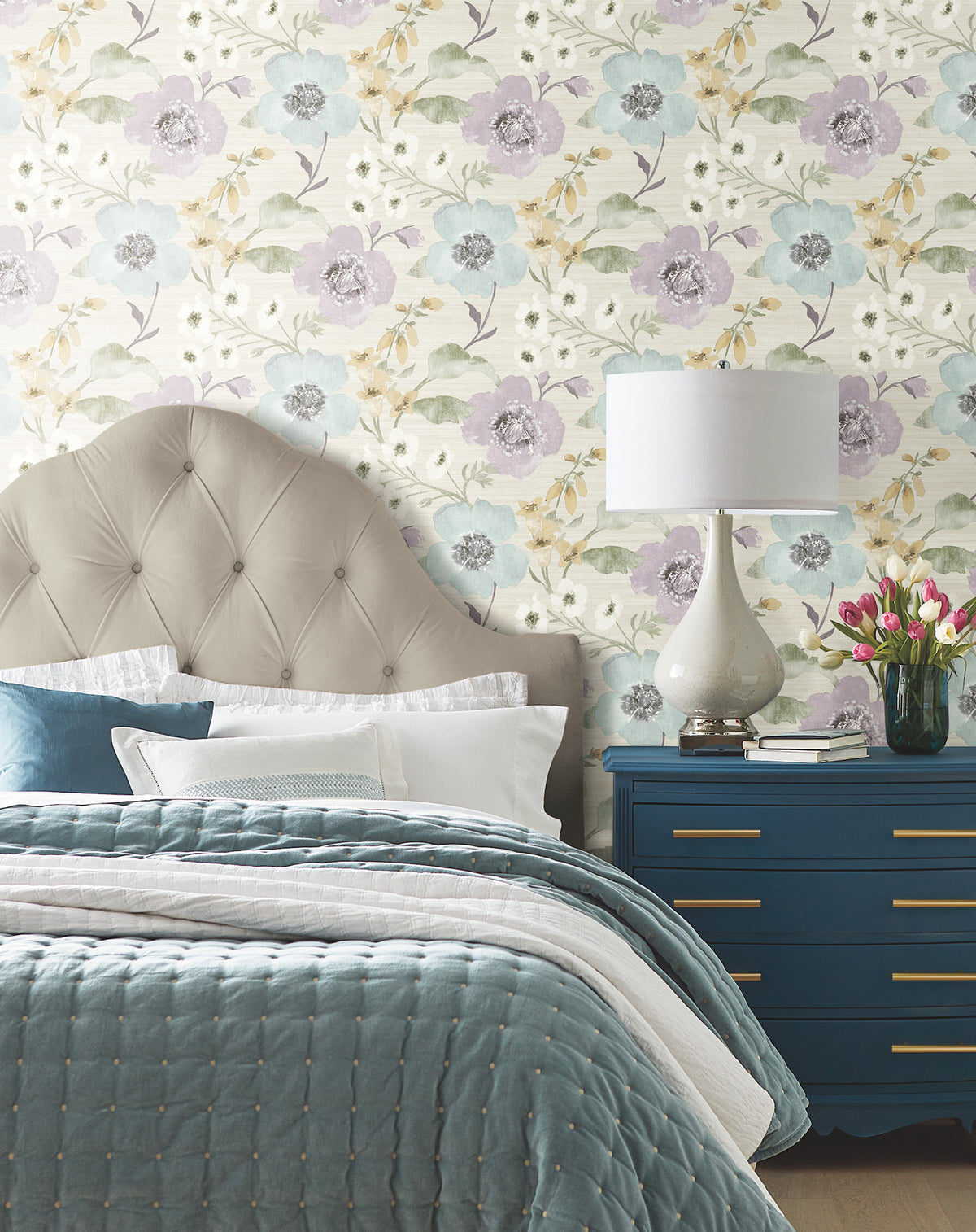 A cozy bedroom featuring a tufted beige headboard, blue quilted bedding, white lamp, a blue dresser with gold handles, and a vase of tulips, set against York Wallcoverings’ Evelyn Floral Wallpaper.