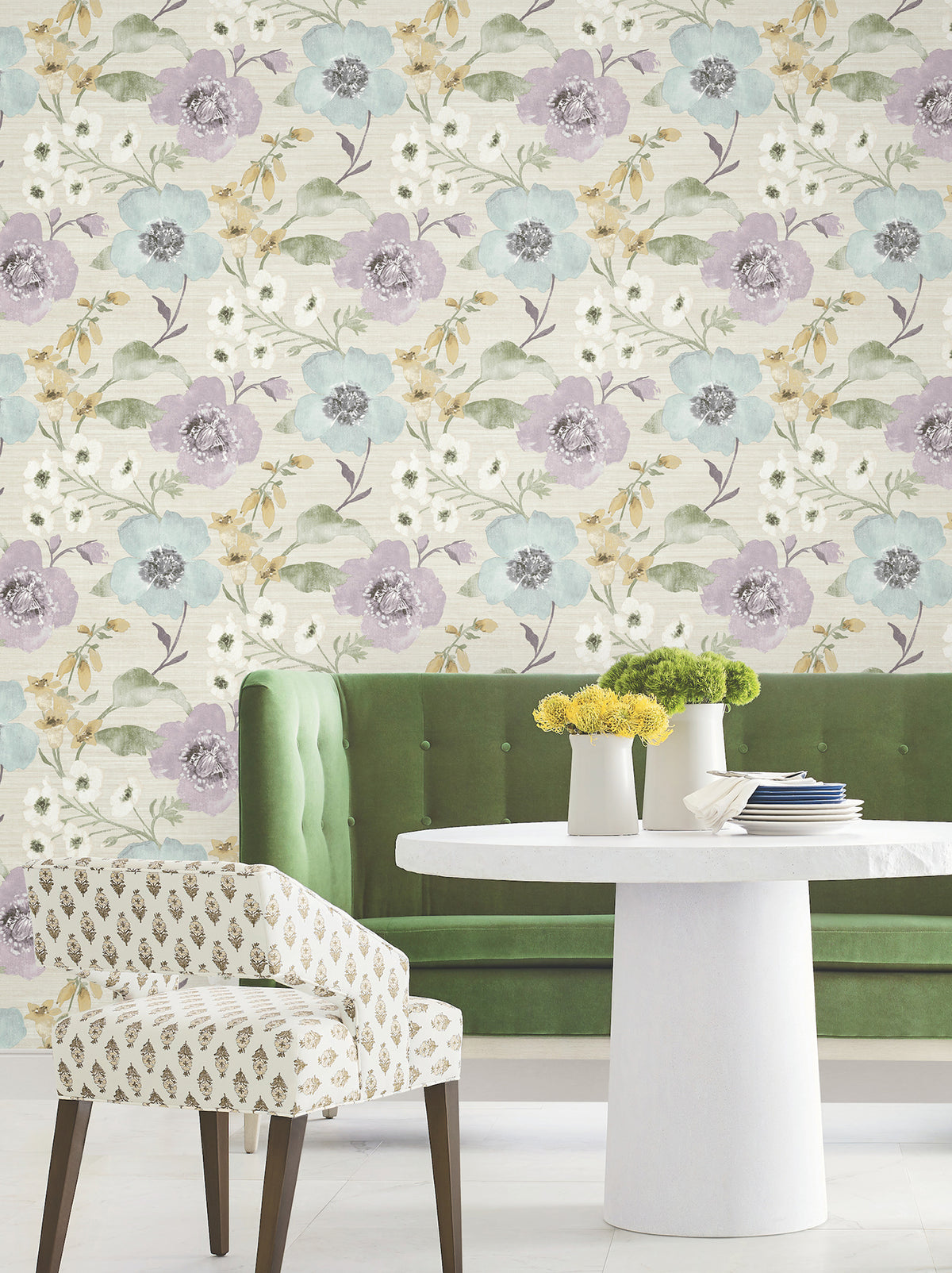 A green velvet bench and a white round table topped with yellow flowers sit in front of York Wallcoverings’ Evelyn Floral Wallpaper. A patterned white chair beside the table completes the cozy, stylish dining space.