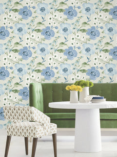 A round white table adorned with yellow and green flowers, a green velvet bench, and a patterned chair are arranged in front of York Wallcoverings’ Evelyn Floral Wallpaper.