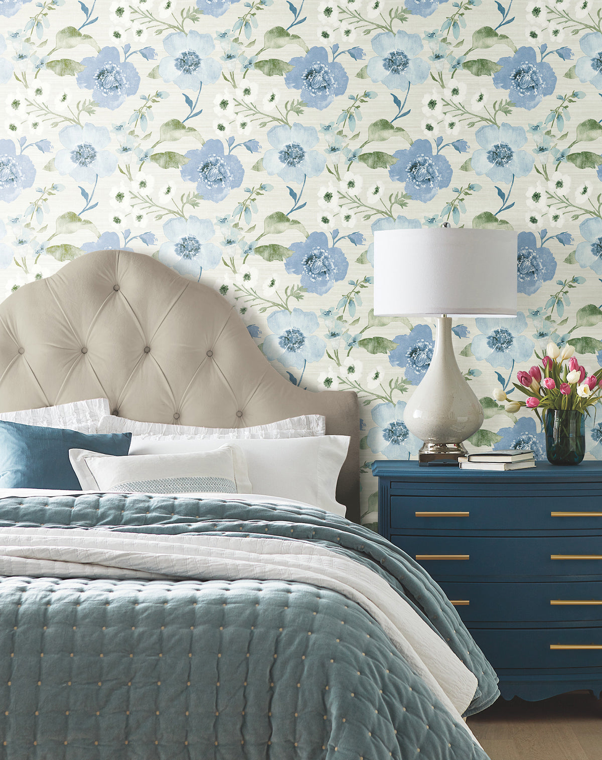 Bedroom featuring Evelyn Floral Wallpaper by York Wallcoverings, a tufted beige headboard, blue quilted bedding, blue dresser with gold handles, white lamp, and a vase of pink tulips. The bed is adorned with white pillows and a blue cushion.
