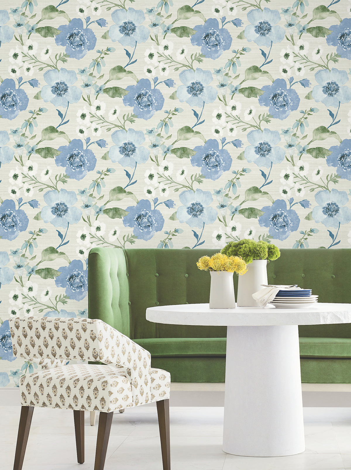 A green tufted bench, round white table, and patterned chair sit in front of a wall covered in Evelyn Floral Wallpaper by York Wallcoverings. Yellow flowers in white vases are arranged on the table.