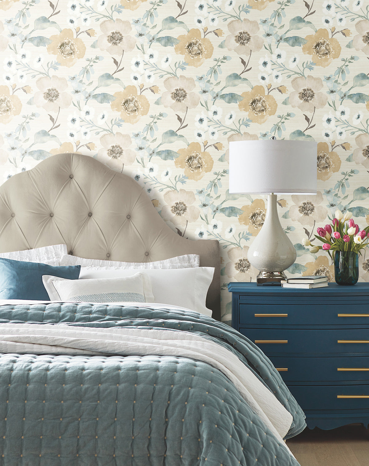 A bedroom featuring a tufted headboard, blue bedding, a blue dresser with gold handles, a white lamp, and a vase of tulips. The wall is adorned with Evelyn Floral Wallpaper by York Wallcoverings, showcasing yellow and blue flowers.