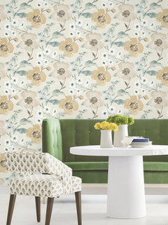 A green tufted bench, a white round table topped with vases of yellow flowers, and a patterned chair are arranged in front of the Evelyn Floral Wallpaper by York Wallcoverings.