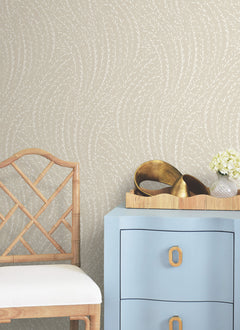 A light wooden chair sits next to a blue drawer with gold handles. On the drawer is a tray with gold decorative objects and a small bouquet of white flowers. The wall features York Wallcoverings’ Willow Song Botanical Wallpaper, adding a subtle beige leafy pattern to the space.