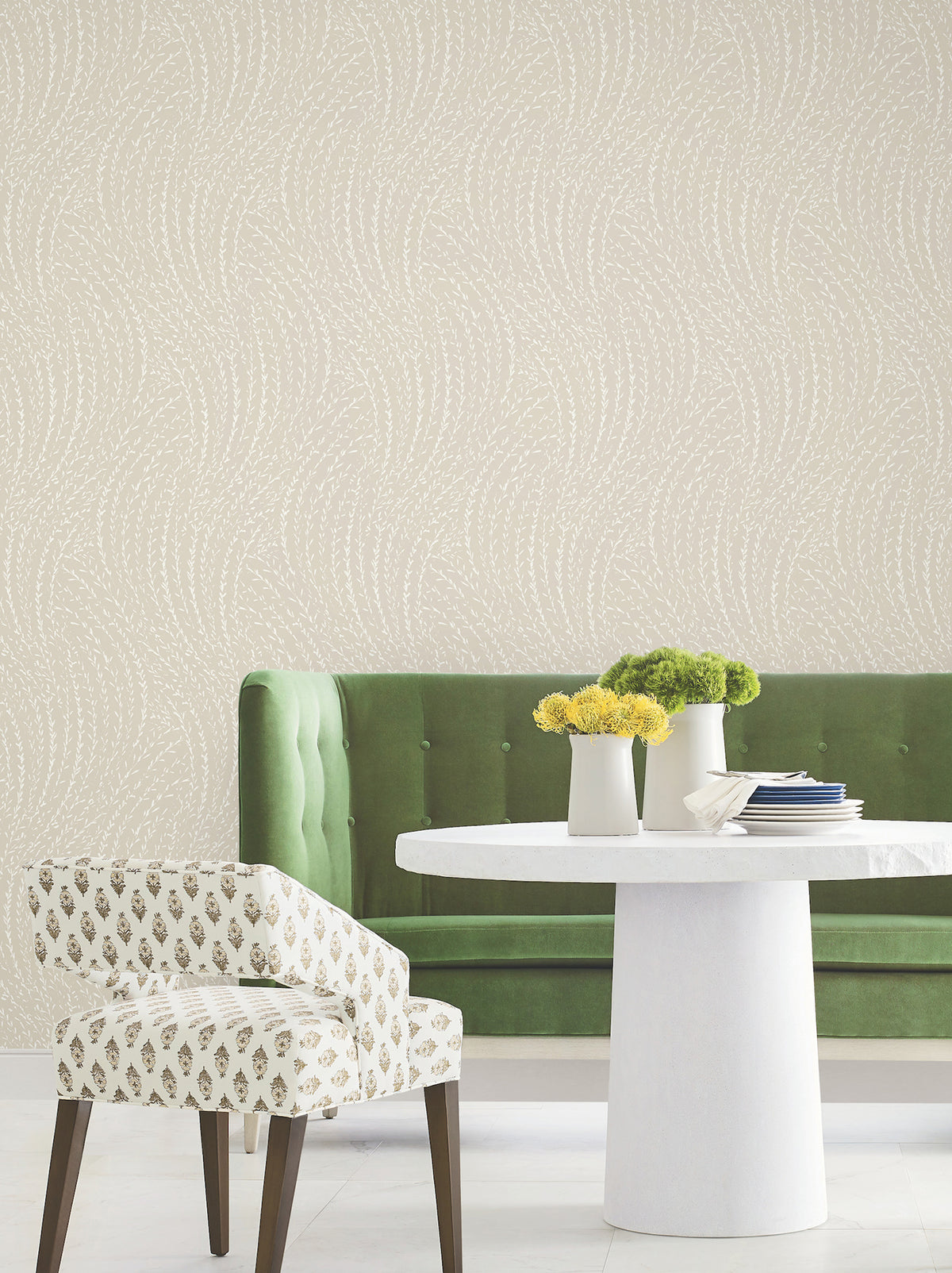 A green velvet sofa, a white patterned chair, and a round white table with yellow flowers are arranged against York Wallcoverings’ Willow Song Botanical Wallpaper, which features a subtle wavy design.