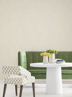 A green velvet sofa, a white patterned chair, and a round white table with yellow flowers are arranged against York Wallcoverings’ Willow Song Botanical Wallpaper, which features a subtle wavy design.