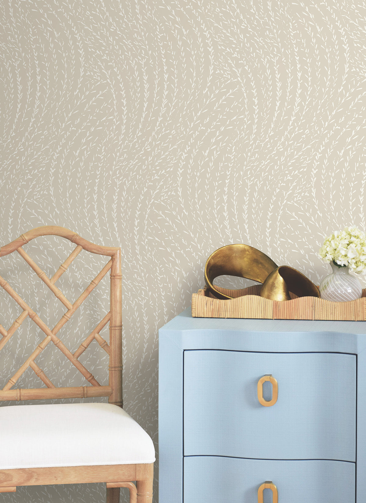 A light blue dresser with gold handles sits next to a wooden chair. On the dresser are a small tray with gold decor and a vase of white flowers, set against York Wallcoverings’ Willow Song Botanical Wallpaper.