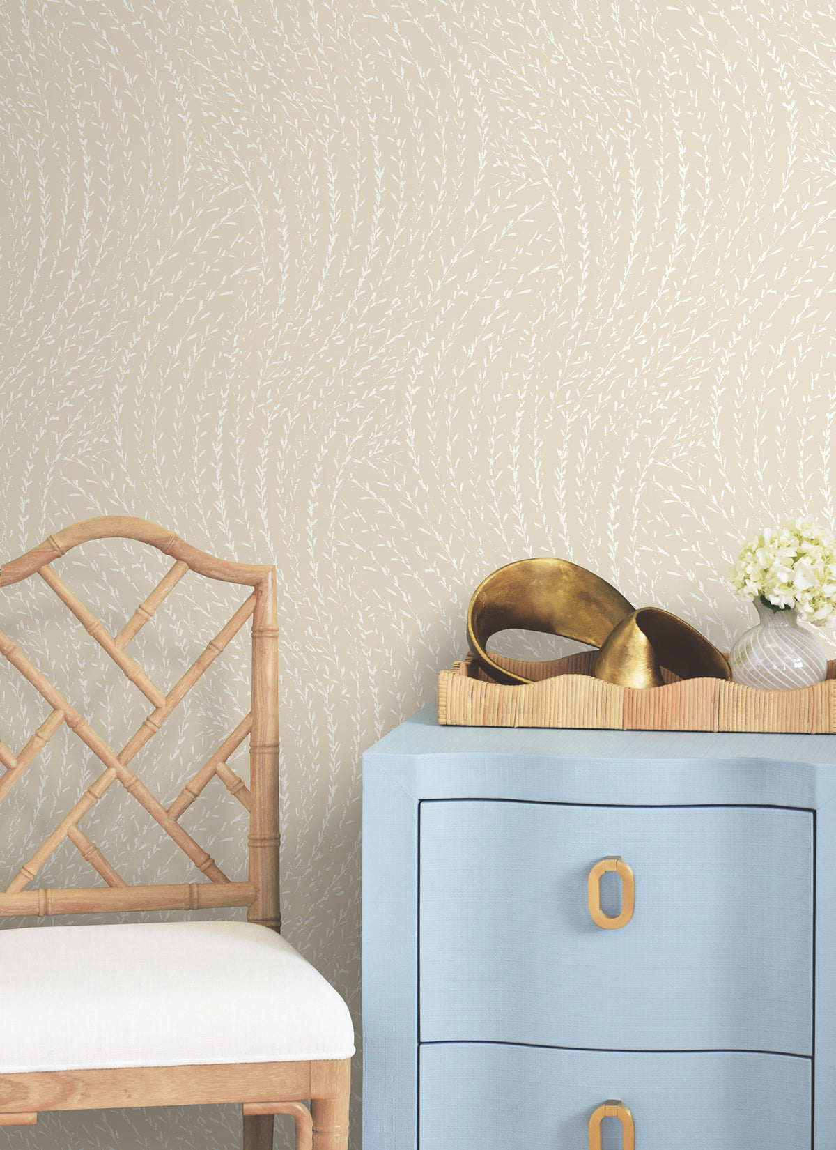 A light wood chair with a white cushion sits beside a blue dresser with gold handles. On top of the dresser are a wooden tray, gold decorative items, and a small arrangement of white flowers. The background features Willow Song Botanical Wallpaper by York Wallcoverings.