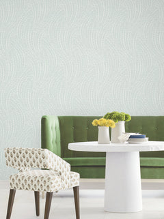 A green velvet tufted bench and a patterned chair sit by a round white table with vases of yellow and green flowers, set against Willow Song Botanical Wallpaper by York Wallcoverings.