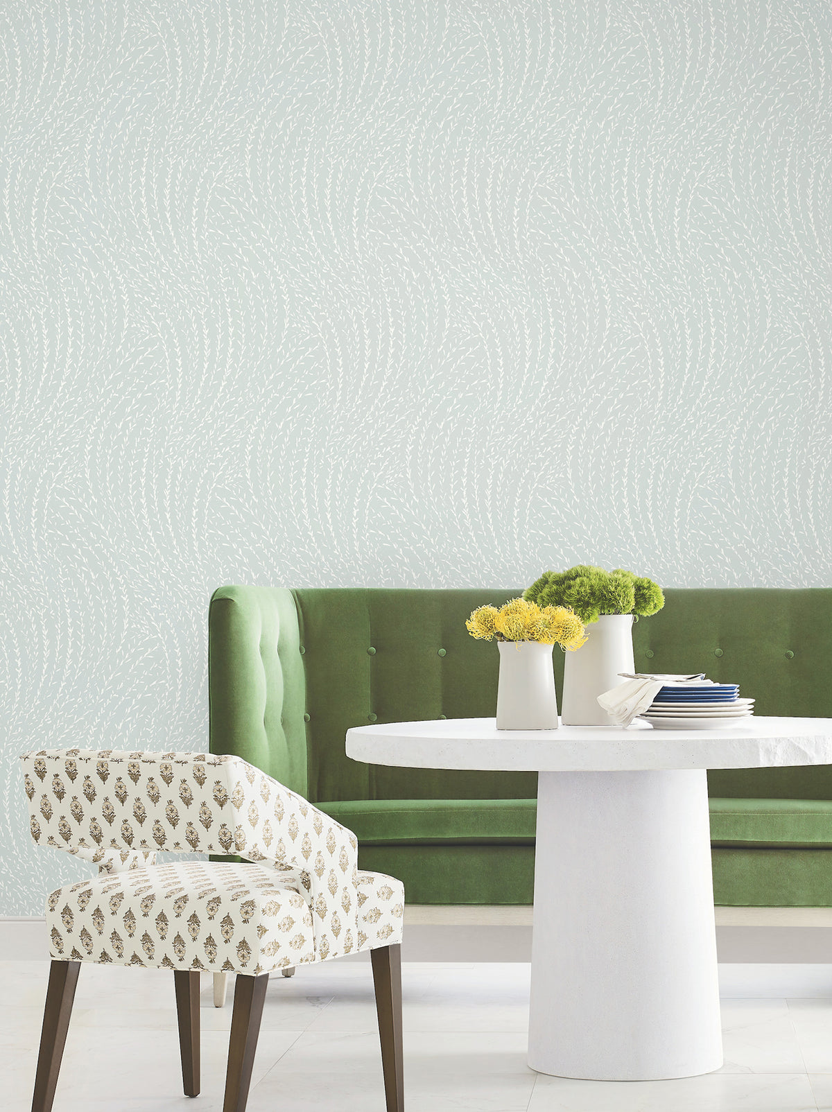 A green velvet bench, a white round table, and a patterned white chair sit in a room with pale Willow Song Botanical Wallpaper by York Wallcoverings. Three small vases with yellow and green flowers are on the table.