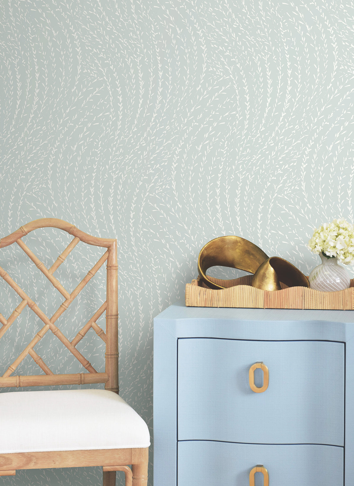 A light blue dresser with brass handles sits next to a bamboo chair with a white cushion. On the dresser are a small bouquet of white flowers and gold decorative bowls. The walls are adorned with Willow Song Botanical Wallpaper by York Wallcoverings, featuring a pale blue background and white leaf pattern.