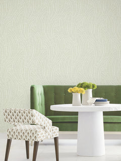 A green tufted bench and a patterned chair are positioned around a round white table adorned with vases of yellow and green flowers, set against Willow Song Botanical Wallpaper by York Wallcoverings, which features a subtle swirling dotted pattern.