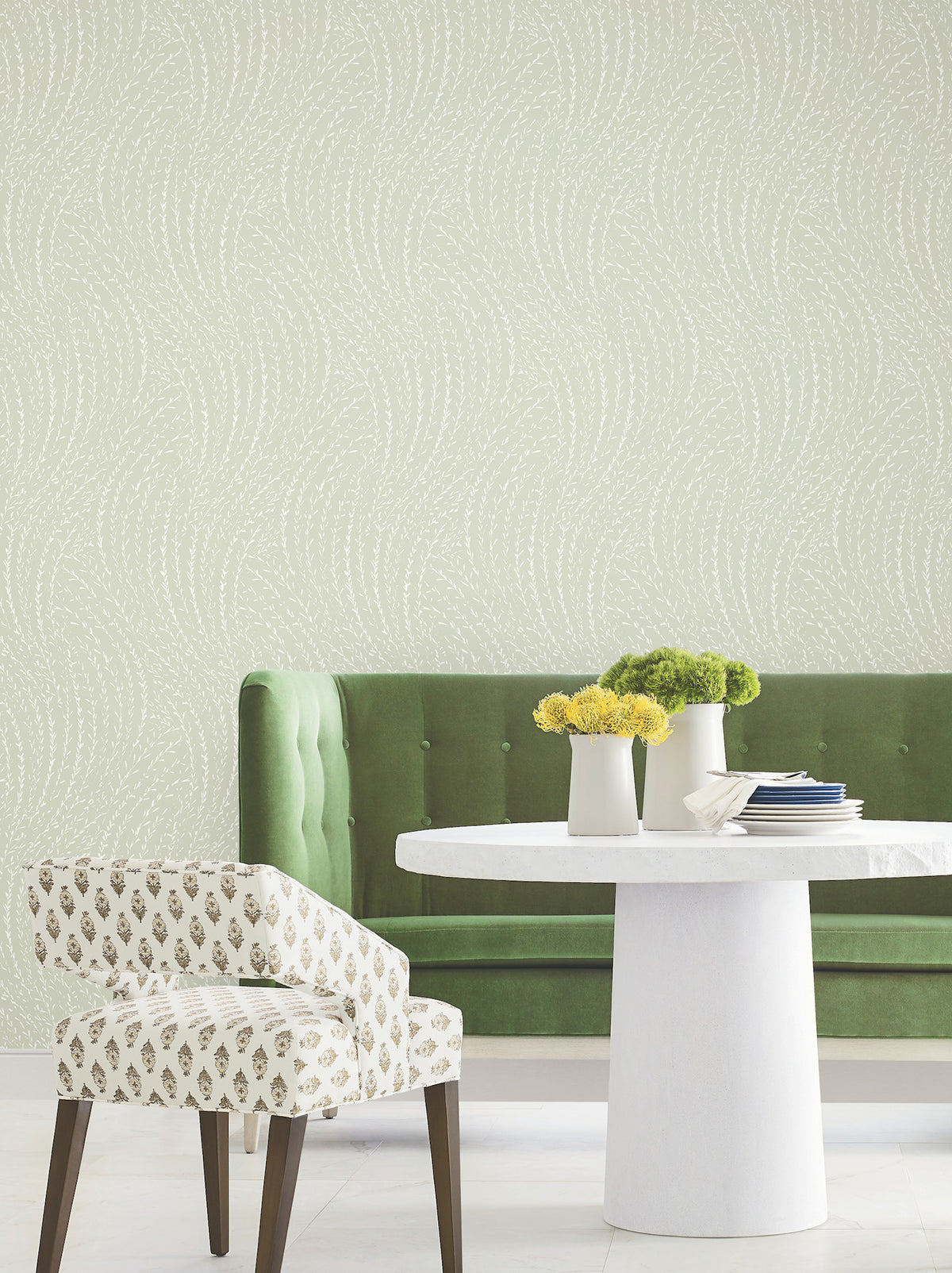 A modern dining area with a green tufted bench, white round table, patterned chair, and York Wallcoverings’ Willow Song Botanical Wallpaper, which features a subtle wavy dotted design. Vases with yellow and green flowers decorate the table.