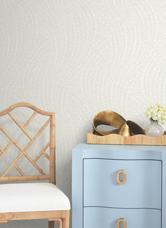 A wooden chair with a white cushion is placed beside a blue drawer. On top of the drawer sits a tray adorned with golden decorative items and a small bouquet of white flowers. The wall behind features the Willow Song Botanical Wallpaper by York Wallcoverings, showcasing its subtle, light-patterned design.