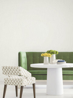 A green velvet sofa, round white table, and patterned chair are arranged in a bright room featuring the Willow Song Botanical Wallpaper by York Wallcoverings with its light, wavy motif. Three white vases holding yellow and green flowers decorate the table.