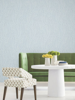 A green velvet tufted bench, a white round table adorned with vases of yellow flowers, and a patterned chair are arranged in front of a blue wall decorated with York Wallcoverings’ Willow Song Botanical Wallpaper.