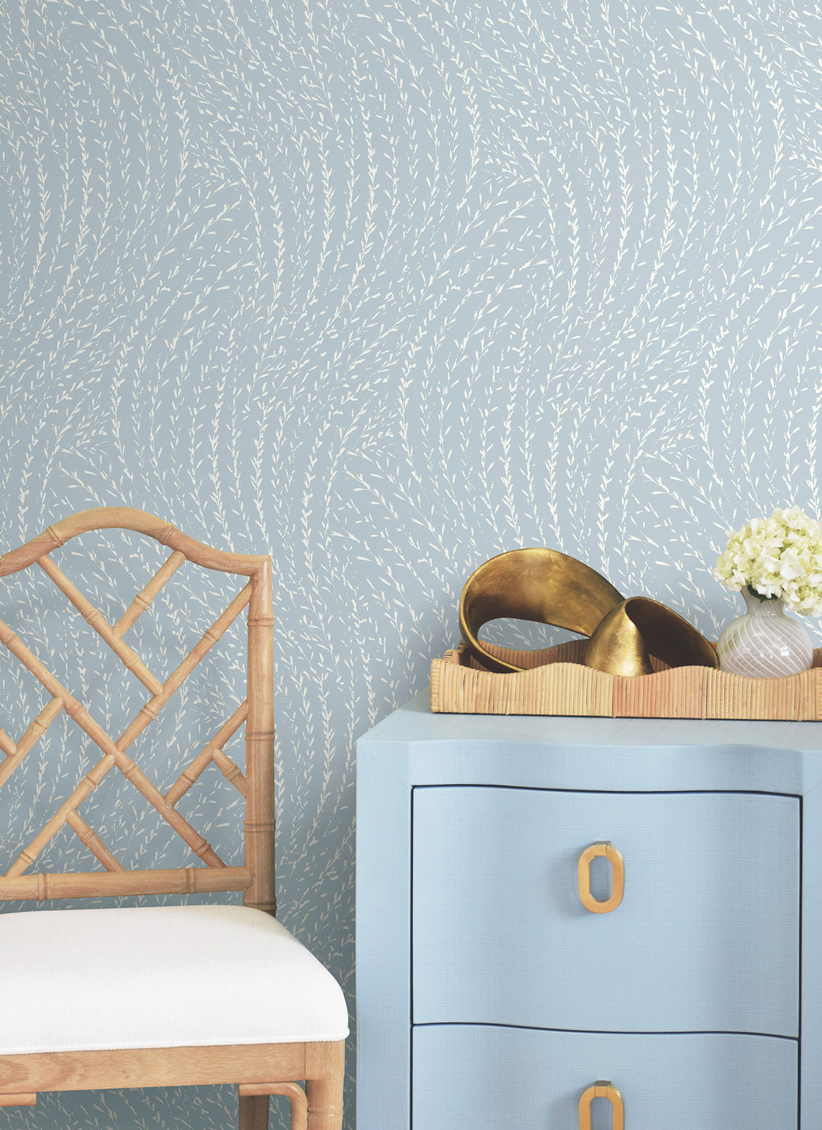 A wall adorned with York Wallcoverings’ Willow Song Botanical Wallpaper, a bamboo chair with a white cushion, a blue dresser with gold handles, a tray featuring gold decor pieces, and a vase of white flowers.