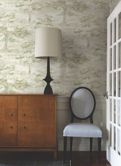 A wooden sideboard with a black lamp, a light lampshade, and a light gray upholstered chair sits against the York Wallcoverings Moreno Woodland Wallpaper, decorated with trees, beside a white French door.