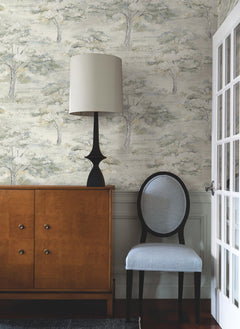 A modern room featuring York Wallcoverings’ Moreno Woodland Wallpaper, a wooden cabinet, a black lamp with a beige shade, and a light blue upholstered chair. A white-paned glass door is on the right.