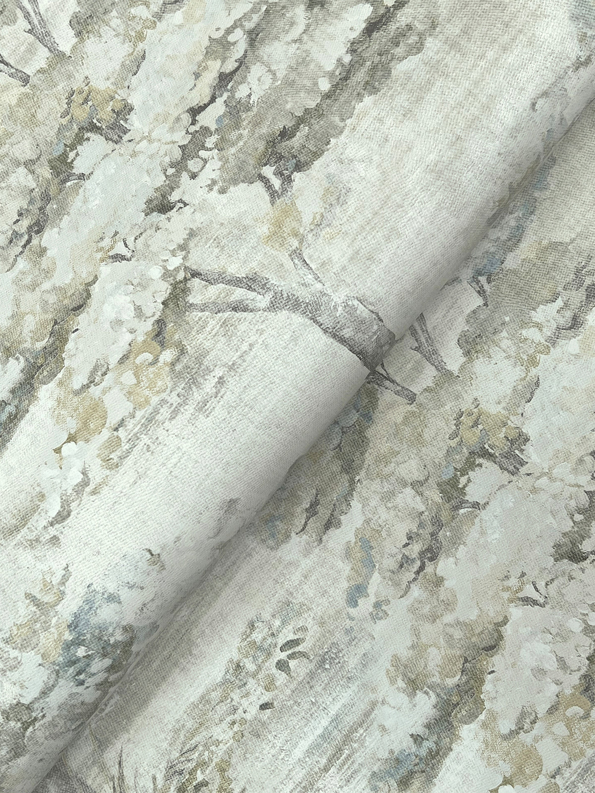 A close-up of Moreno Woodland Wallpaper by York Wallcoverings shows its delicate, muted pattern featuring trees with leafy branches in soft shades of gray, green, and cream. The wallpaper appears slightly folded, creating gentle shadows.