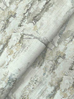 A close-up of Moreno Woodland Wallpaper by York Wallcoverings shows its delicate, muted pattern featuring trees with leafy branches in soft shades of gray, green, and cream. The wallpaper appears slightly folded, creating gentle shadows.