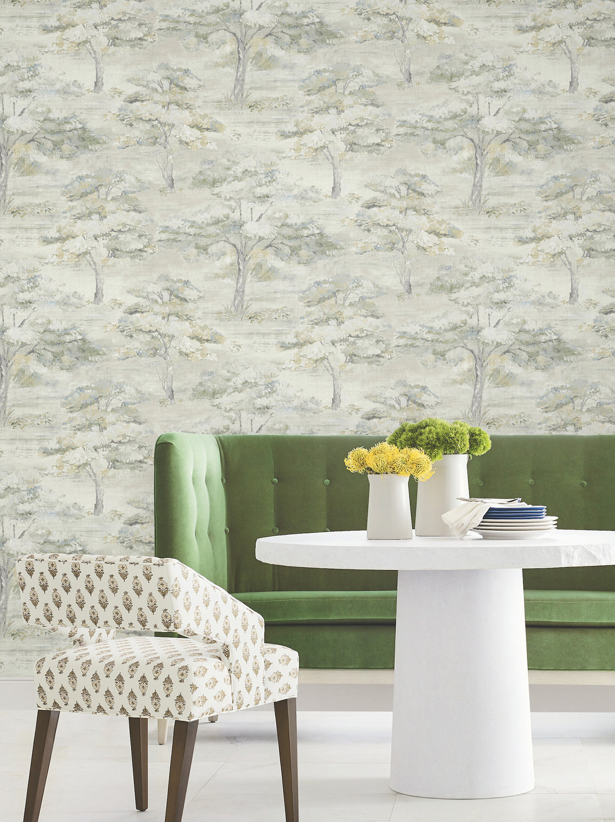 Moreno Woodland Wallpaper by York Wallcoverings featuring a light green and gray tree design provides a bright and modern backdrop for the setting, which includes a green tufted bench, white round table with yellow flowers, and a patterned chair.