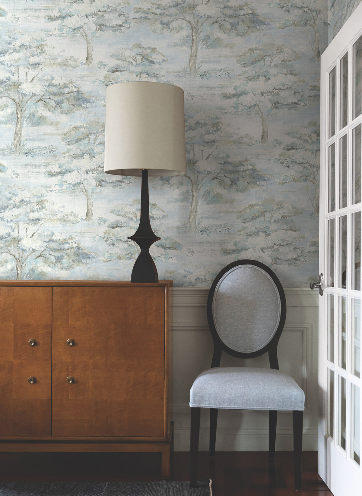 A wooden cabinet with a black lamp and beige shade sits next to a light gray upholstered chair. The wall features York Wallcoverings’ Moreno Woodland Wallpaper, and glass-paned doors are partly visible on the right.