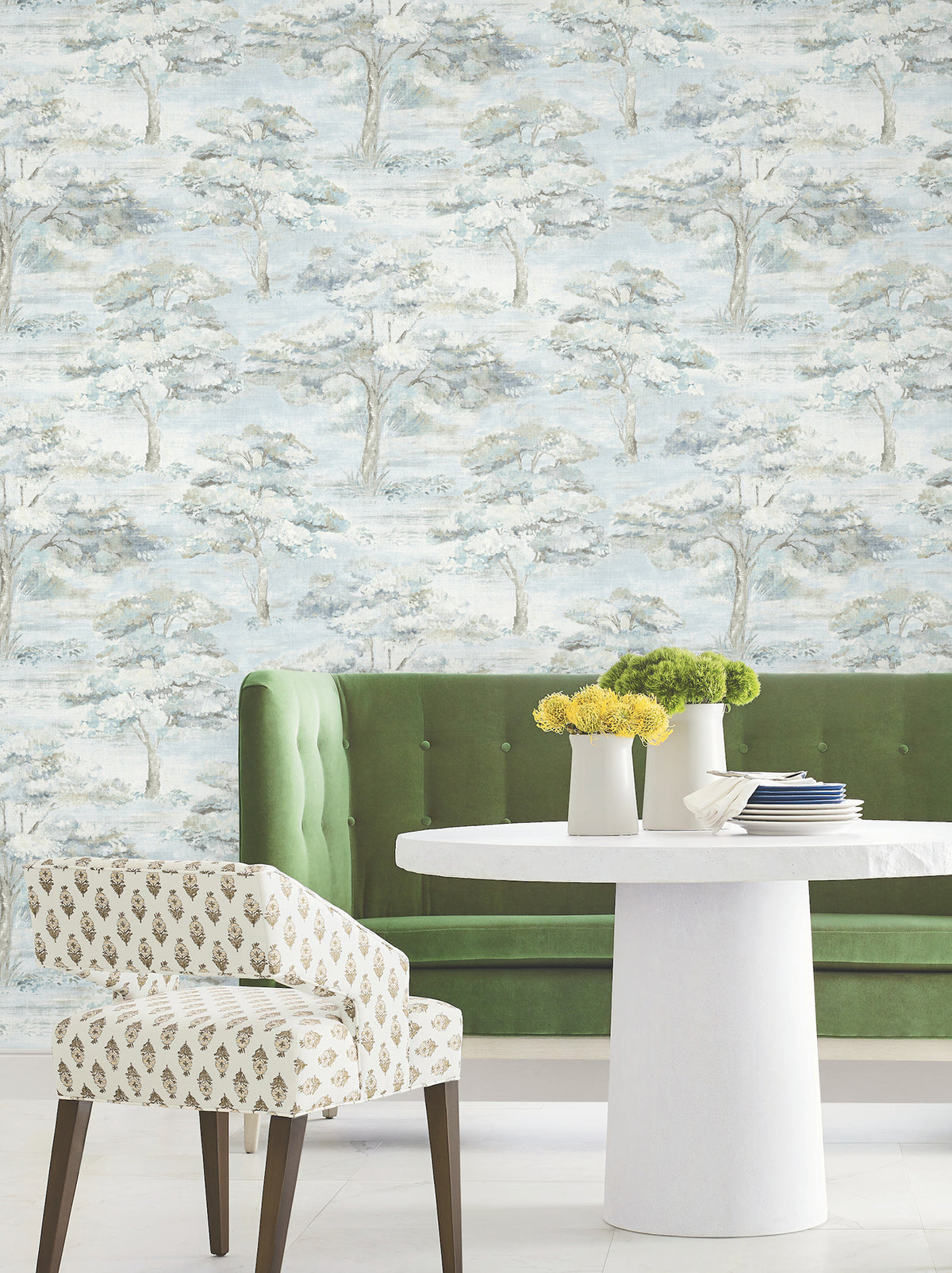 A green tufted sofa, round white table with vases of yellow flowers, and a patterned chair sit in front of a wallpapered wall featuring the Moreno Woodland Wallpaper by York Wallcoverings, which displays a soft blue and white tree landscape.
