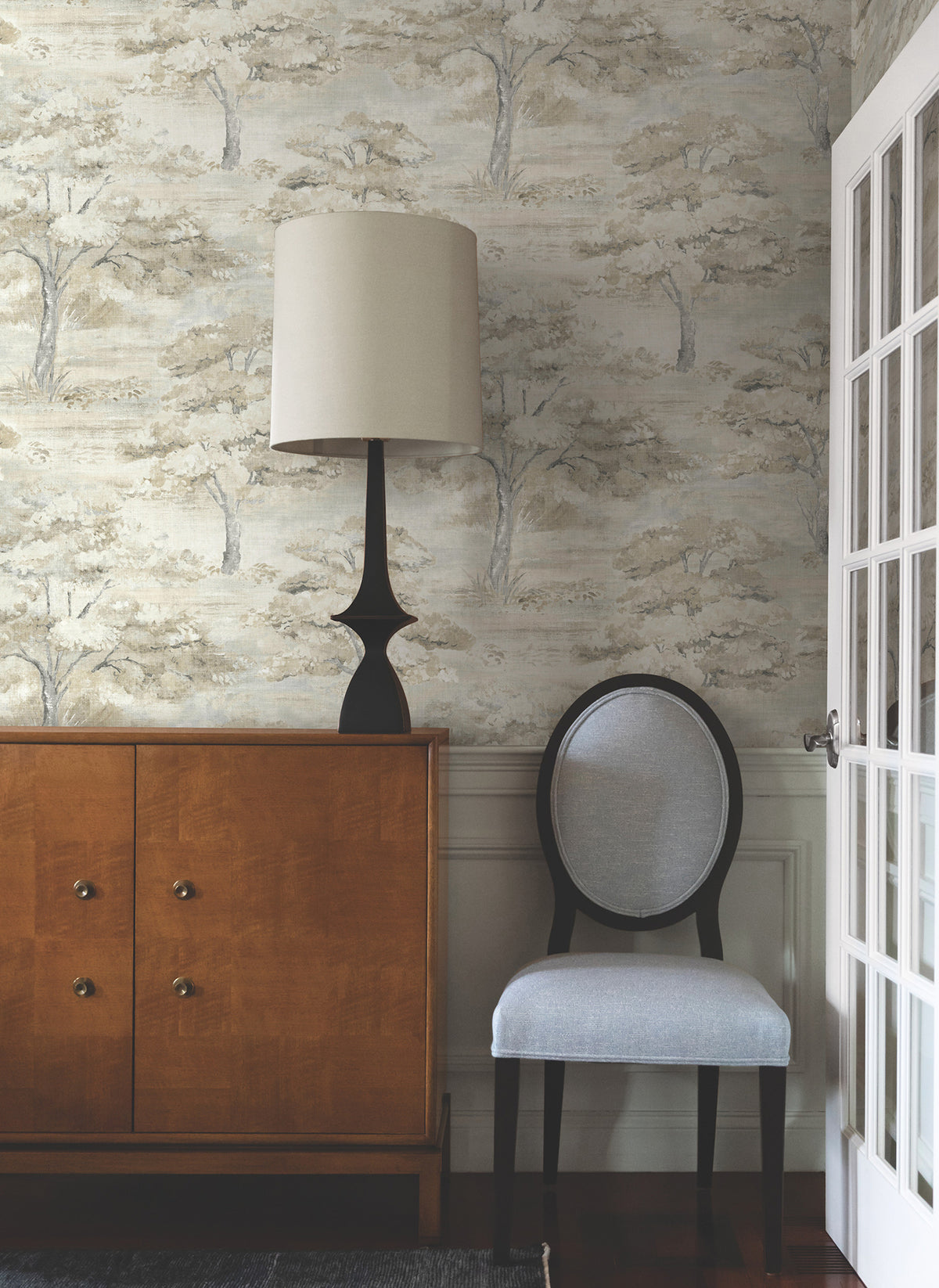 A wooden cabinet with a black lamp sits beside a gray upholstered chair. Behind them, Moreno Woodland Wallpaper by York Wallcoverings adds a scenic tree-patterned backdrop, while a white-paned glass door stands to the right.
